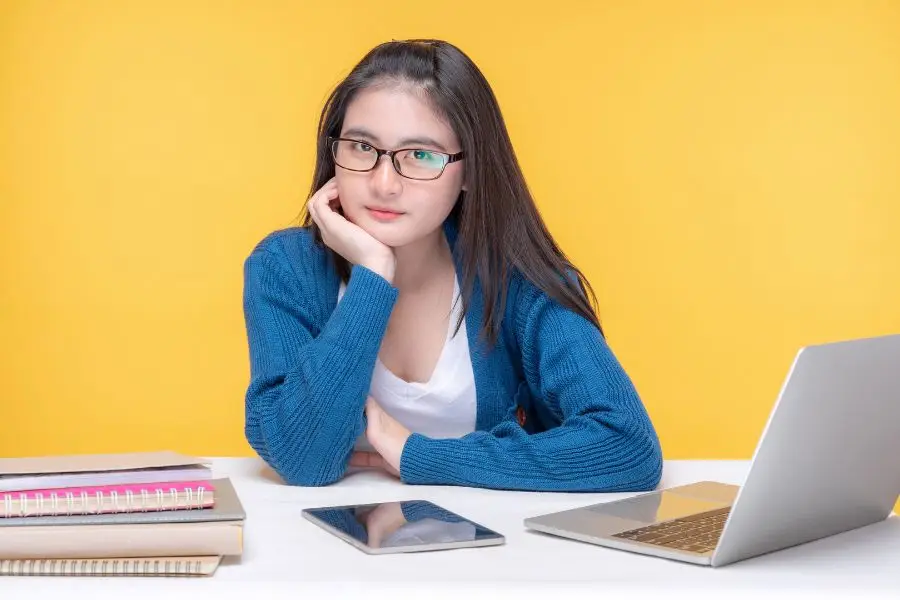A young woman wearing glasses and a blue sweater, sitting at a desk with a laptop and a tablet, looking thoughtfully at the camera against a yellow background.